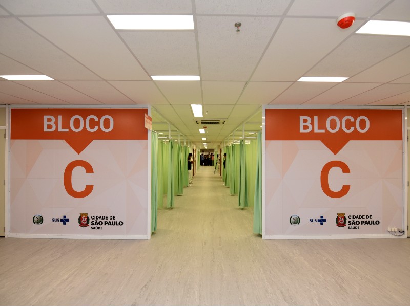Foto interna do Hospital Municipal Lydia Storópoli que mostra um corredor com várias divisórias dos dois lados separadas por cortinas verdes onde ficam os leitos. Na entrada, há duas placas grandes, uma de cada lado do corredor, onde está escrito Bloco C na cor laranja